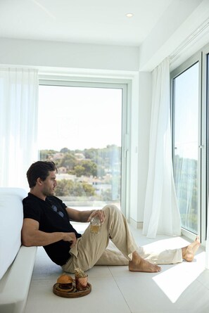 A man sitting on a white sofa by a large window with a city view, holding a glass of drink and a burger on a coaster.