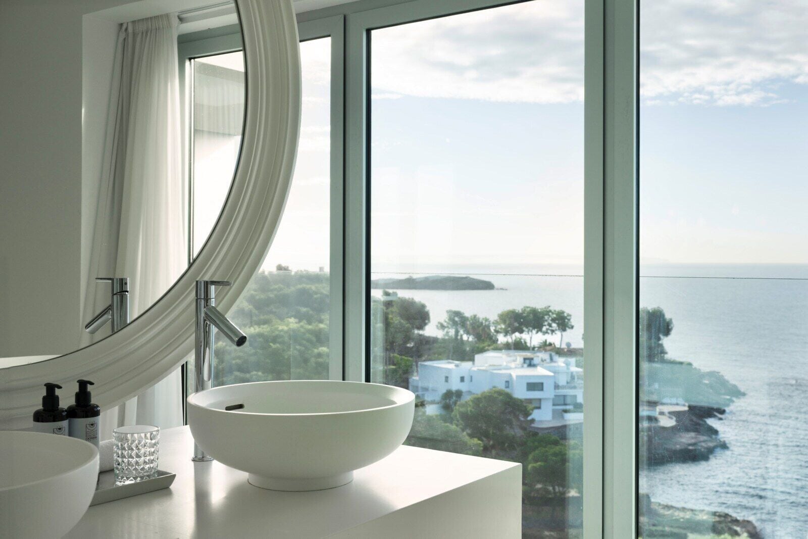Un baño moderno con un ventanal grande, un lavabo blanco y vistas al mar y a edificios.