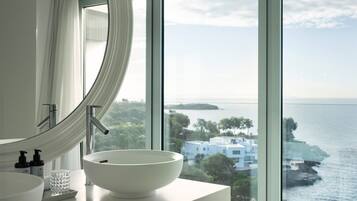 Un baño moderno con un ventanal grande, un lavabo blanco y vistas al mar y a edificios.