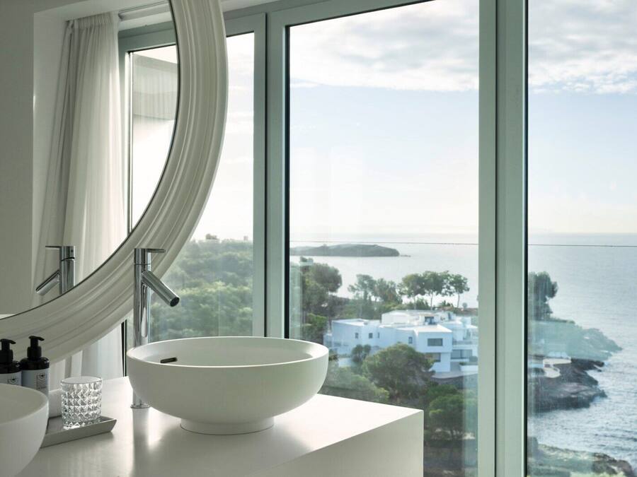 Un baño moderno con un ventanal grande, un lavabo blanco y vistas al mar y a edificios.