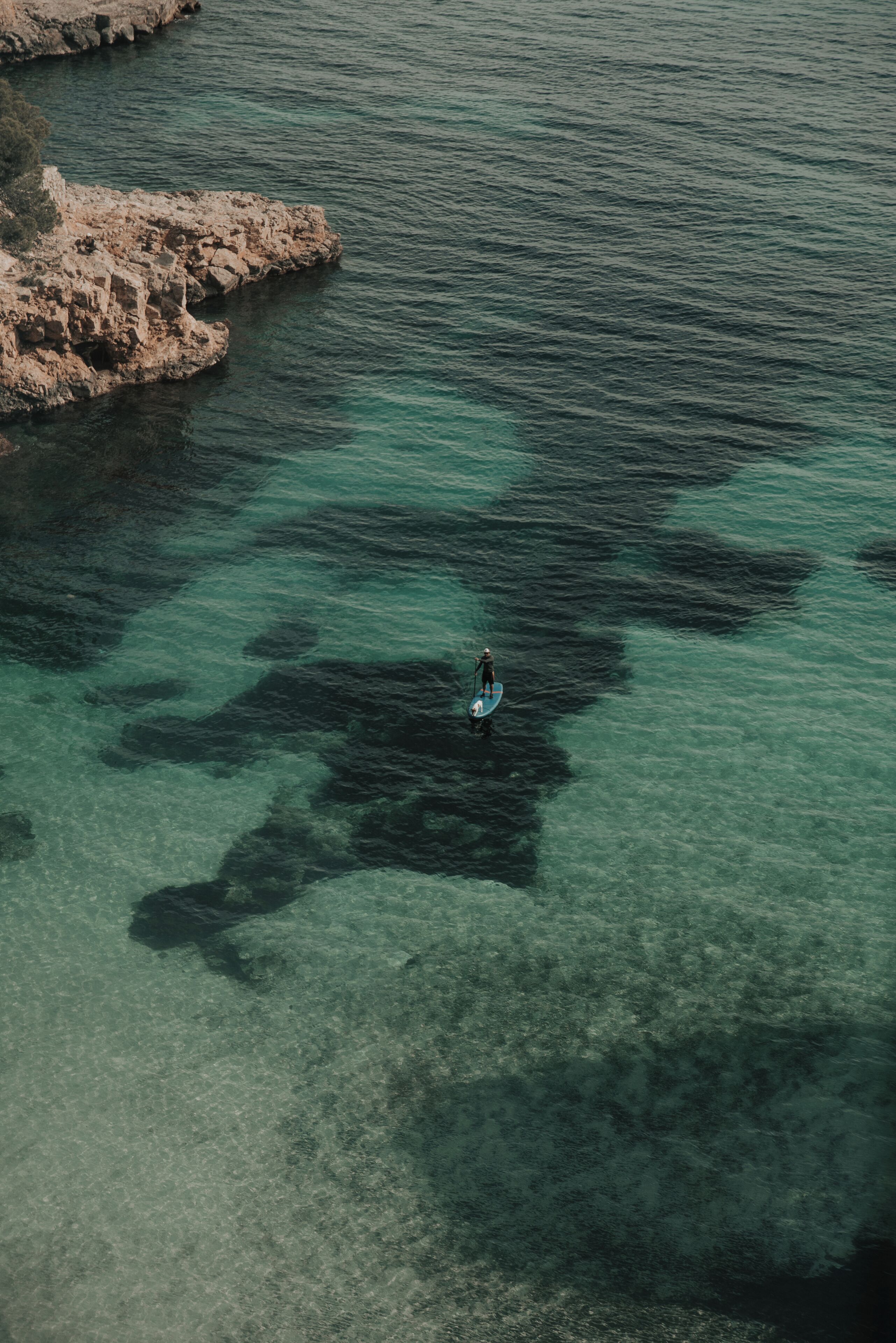 A person paddleboarding in clear turquoise waters near rocky terrain.
