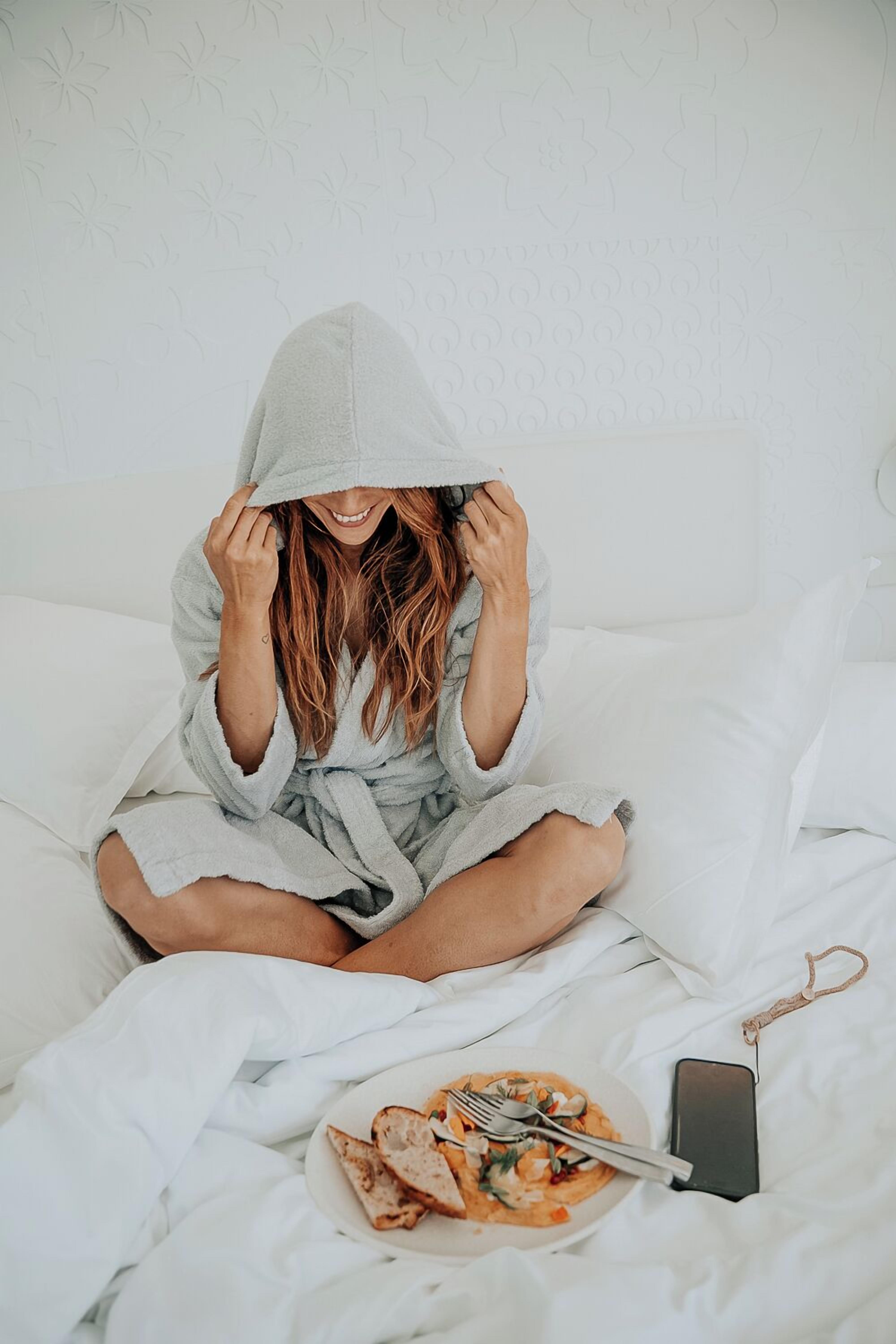 Uma pessoa sentada na cama com um prato de comida e um smartphone ao lado.