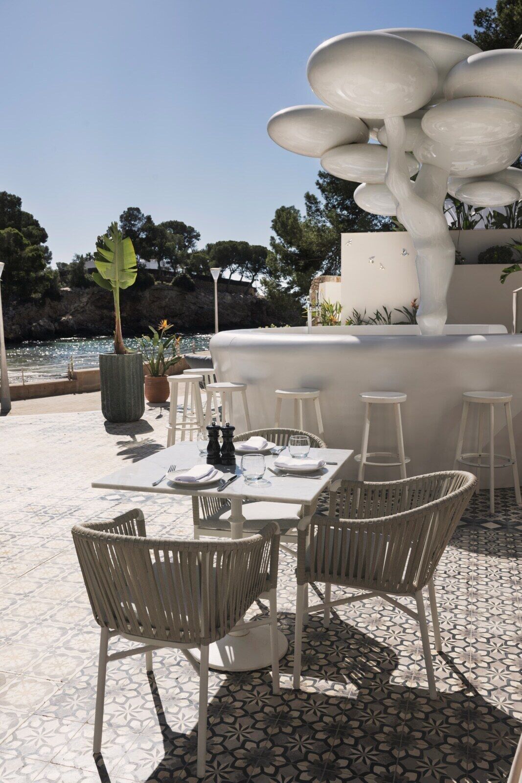 Un área de comedor al aire libre con sillas y mesas de mimbre blanco, una fuente decorativa blanca y vista al mar.