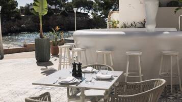 Un área de comedor al aire libre con sillas y mesas de mimbre blanco, una fuente decorativa blanca y vista al mar.
