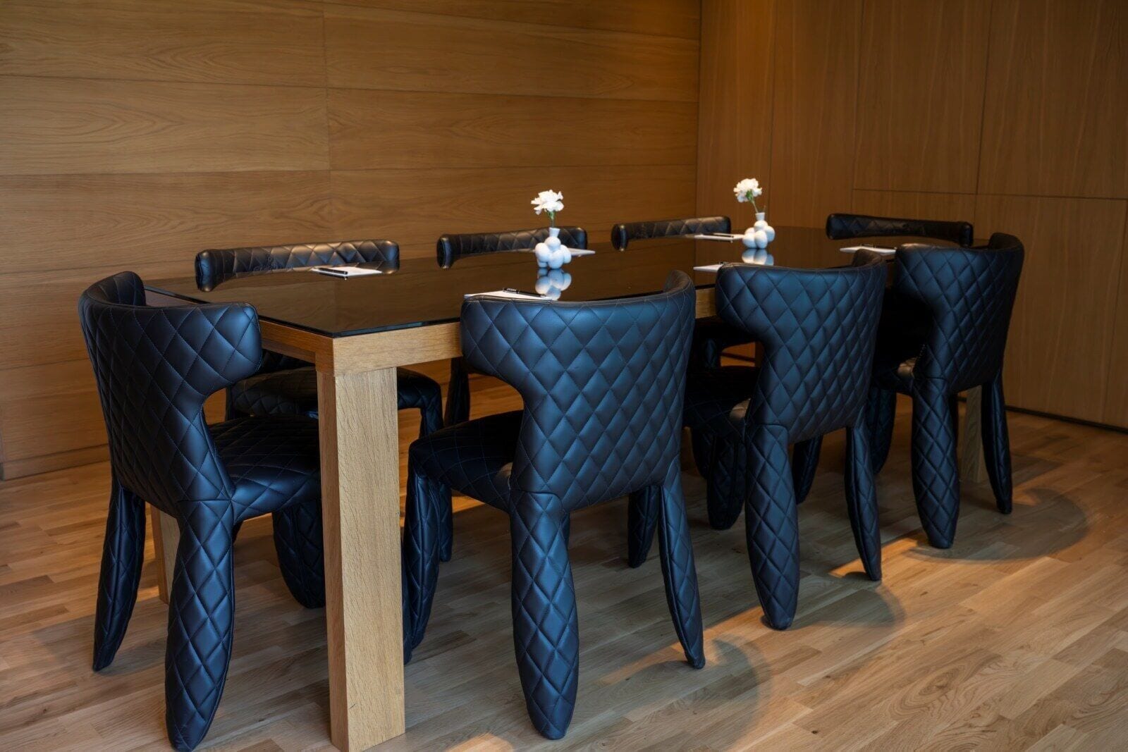 A conference table with six black leather chairs, a wooden table, and white vases with flowers.