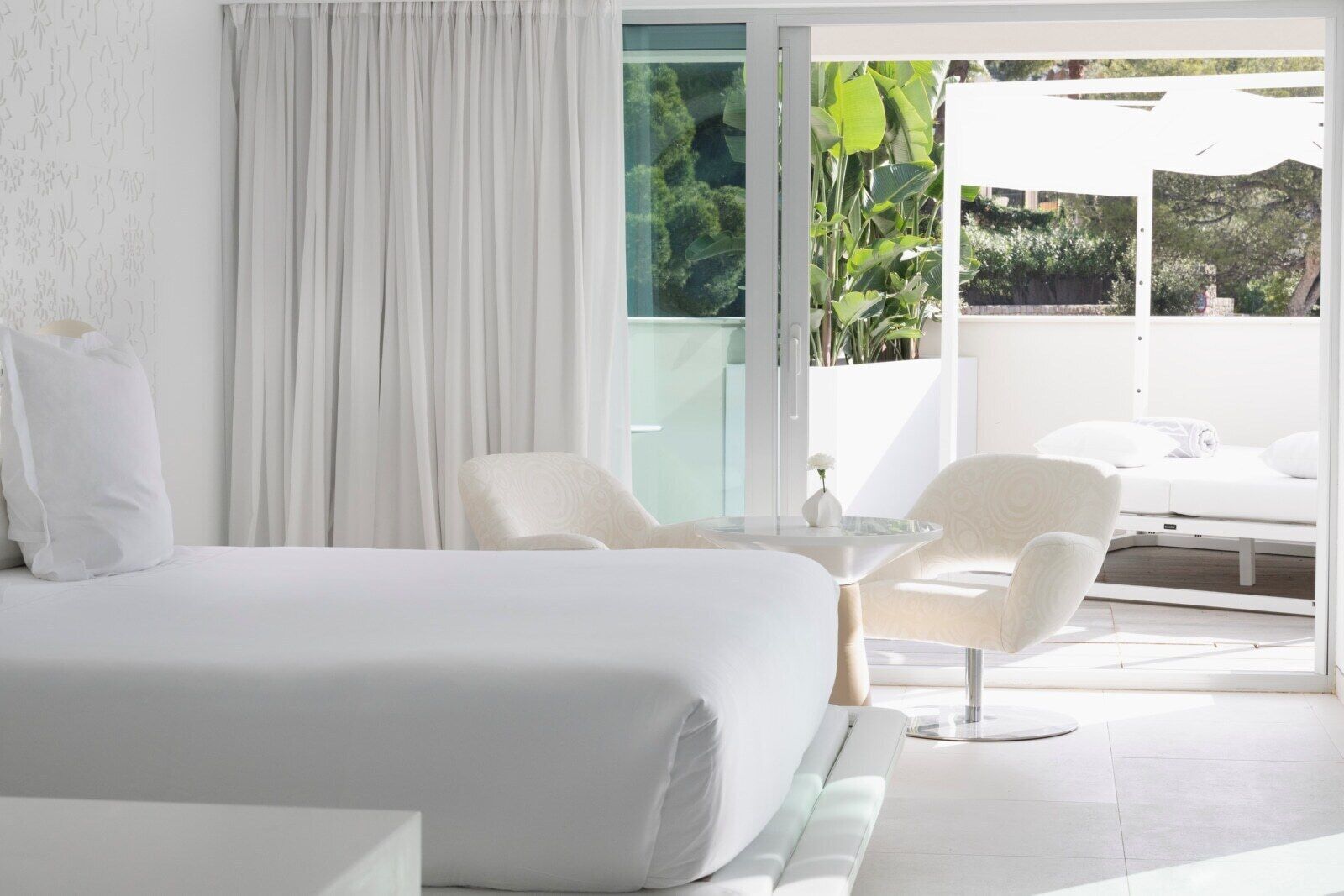 A modern hotel room with a large bed, a small table, two chairs, and a balcony with a view of greenery.