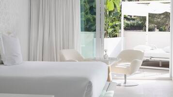 A modern hotel room with a large bed, a small table, two chairs, and a balcony with a view of greenery.