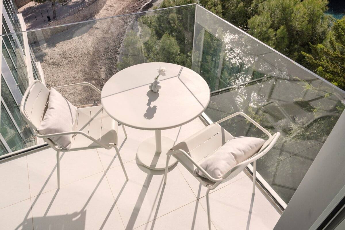 A balcony with a glass railing, a round table, and two white chairs with cushions.