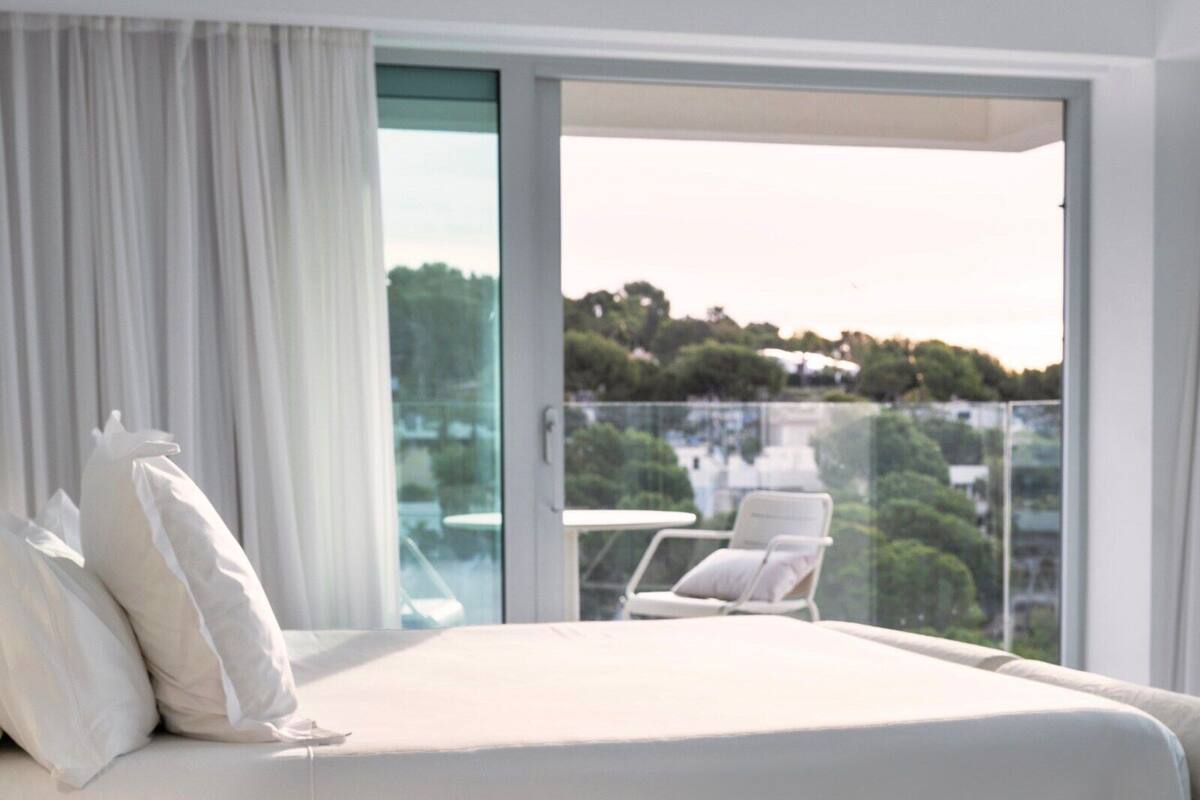 A hotel room with a bed, white curtains, a balcony with a chair, and a view of greenery and buildings.