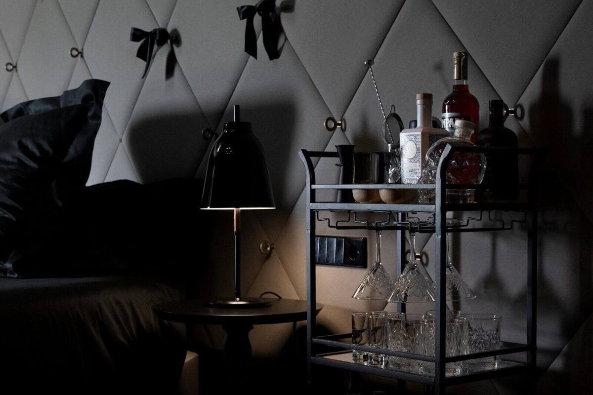 A dimly lit room with a tufted headboard, a black lamp, a glassware rack, and a bottle of liquor.