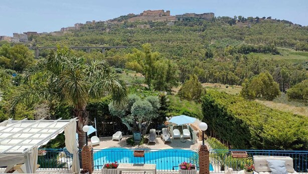 Área de piscina com espreguiçadeiras e guarda-sóis, cercada por vegetação e vista para uma colina com edifícios.