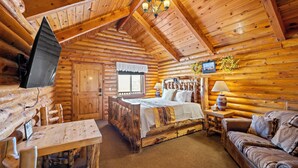Interior de una cabaña de troncos con una cama de madera, una mesa de comedor de madera, un sofá y un televisor montado en la pared.