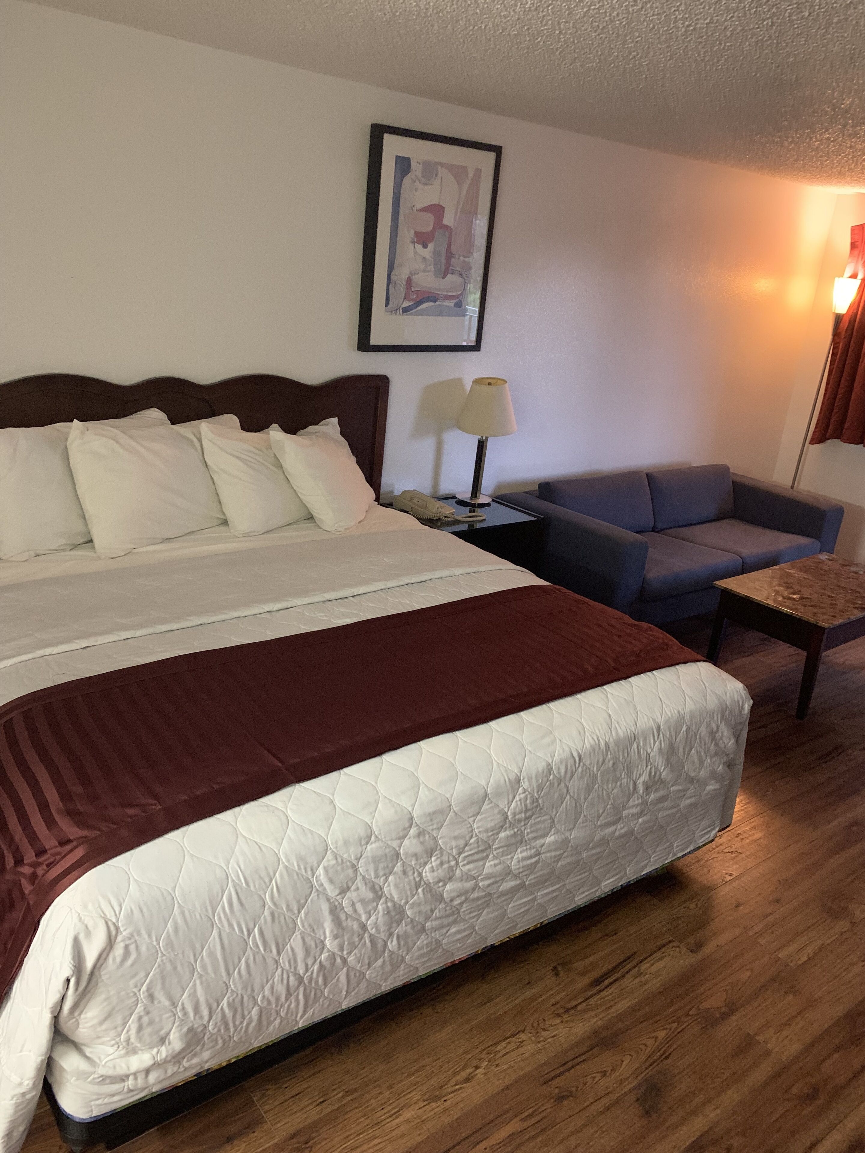 Habitación de hotel con una cama grande, un sofá, una mesita de noche con lámpara y un cuadro en la pared.