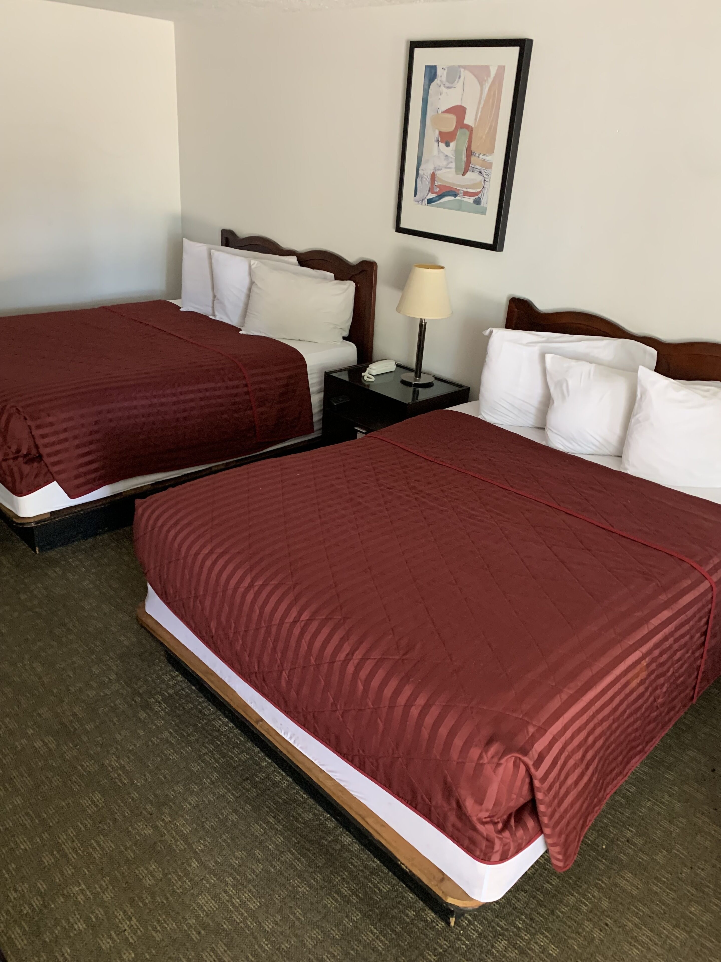 A hotel room with two beds, each with a red bedspread and white pillows. There is a nightstand with a lamp and a phone between the beds. A framed picture hangs on the wall above the beds.