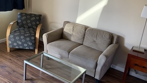 A living room with a beige sofa, a patterned armchair, a glass coffee table, and a lamp.