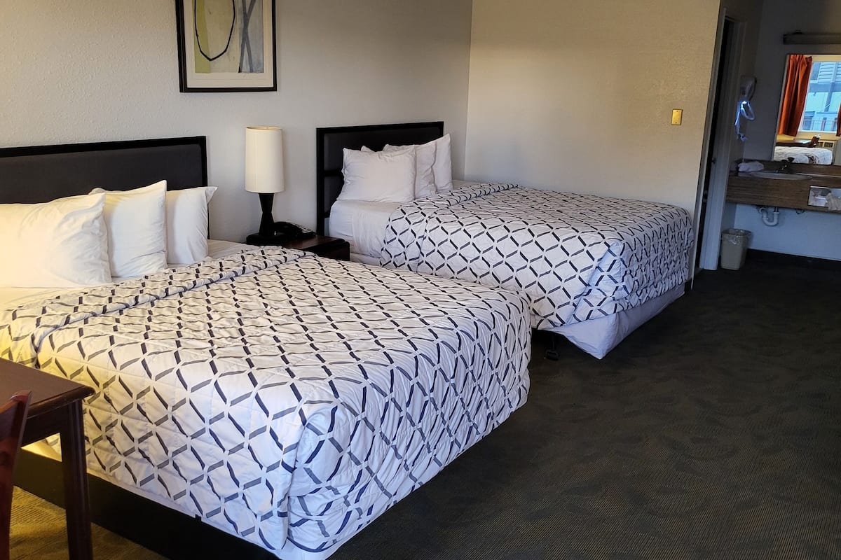 A hotel room with two beds, a desk, a chair, a lamp, and a framed artwork on the wall.