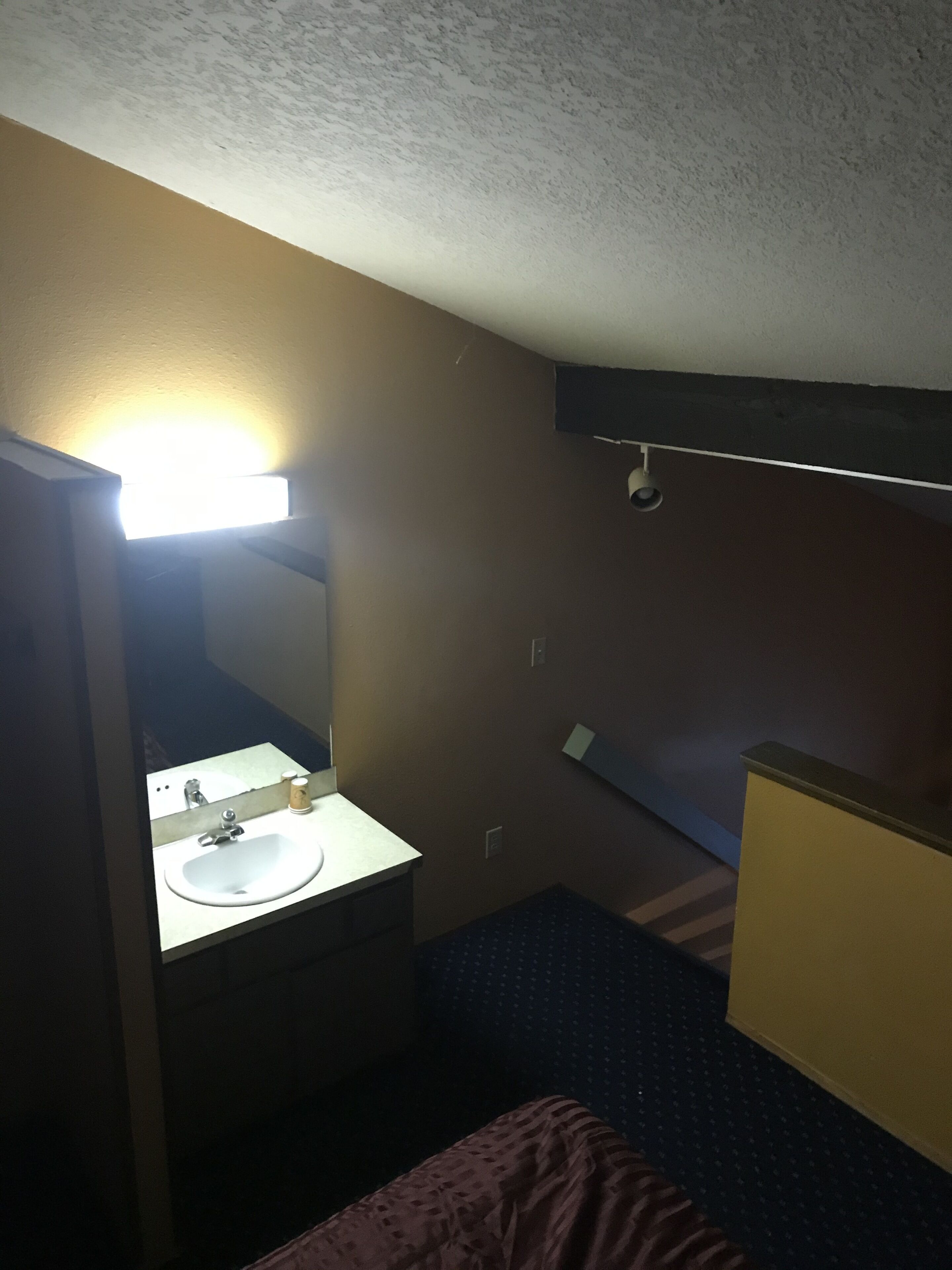 Une petite pièce faiblement éclairée, avec un lit simple, une table de chevet et un lavabo rudimentaire agrémenté d’un miroir.