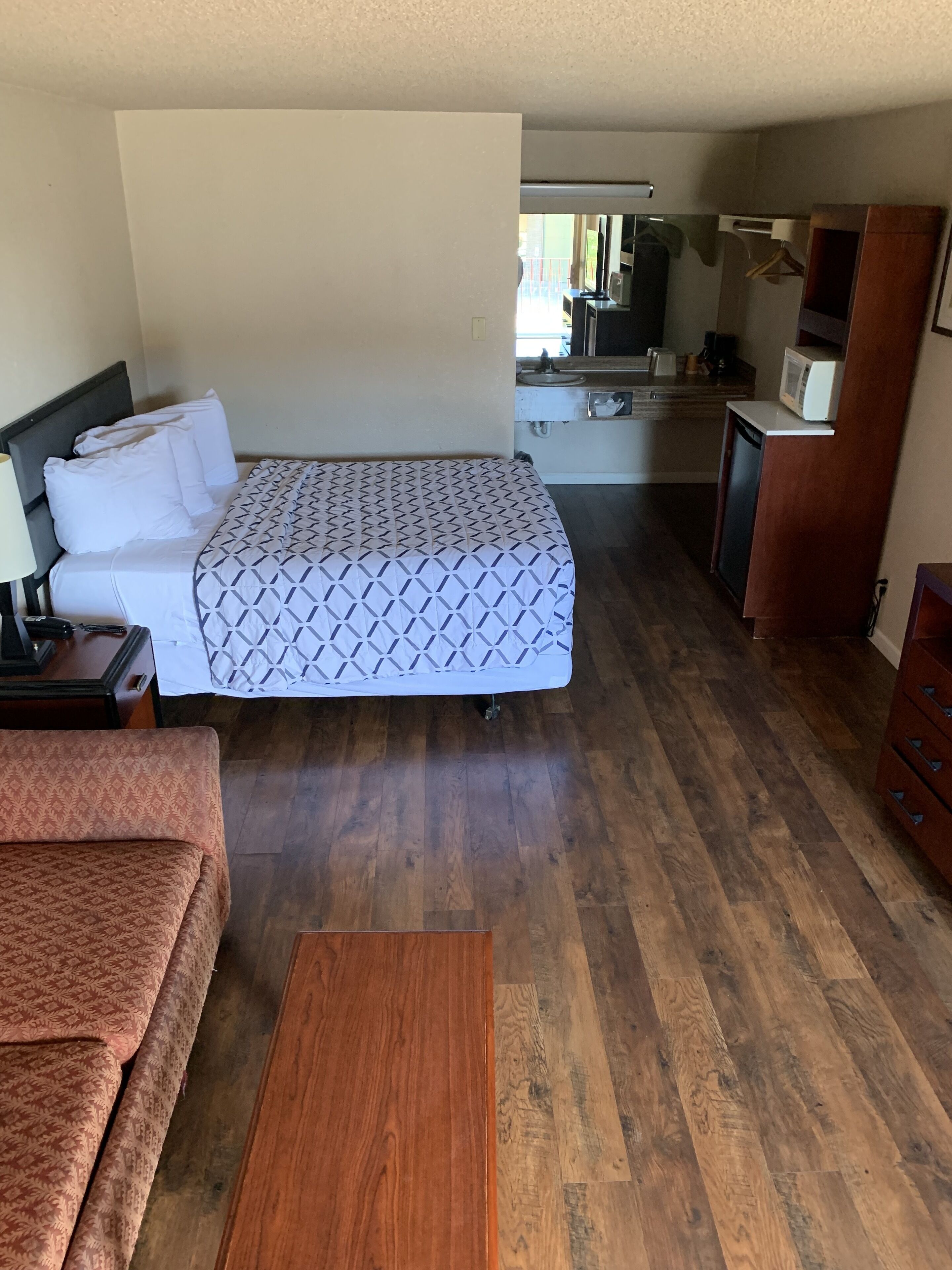 Habitación de hotel compacta con cama, un pequeño sofá, mesa de madera, kitchenette y baño.