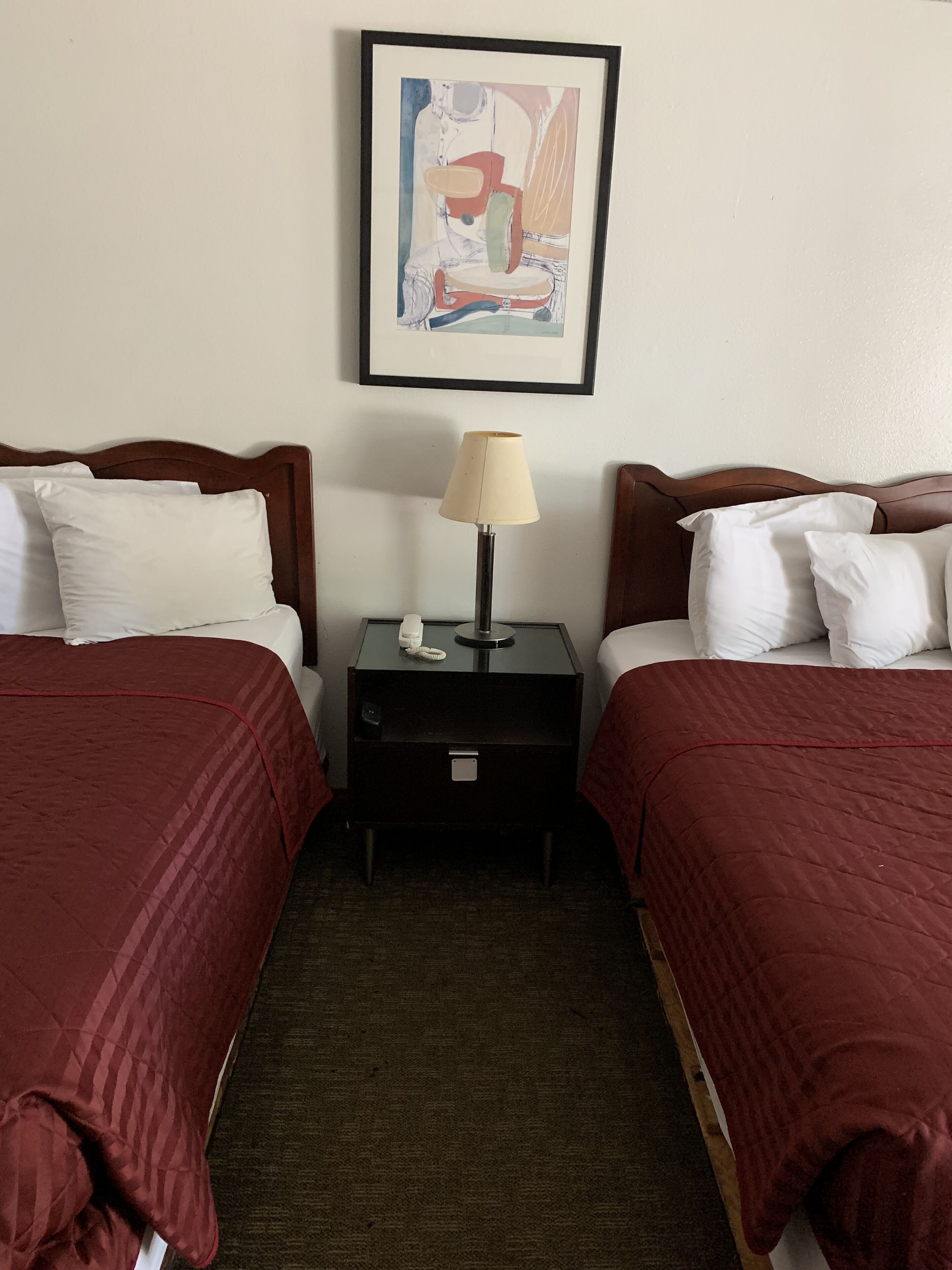 A hotel room with two beds, each with a red bedspread, a nightstand with a lamp, and a framed abstract painting on the wall.