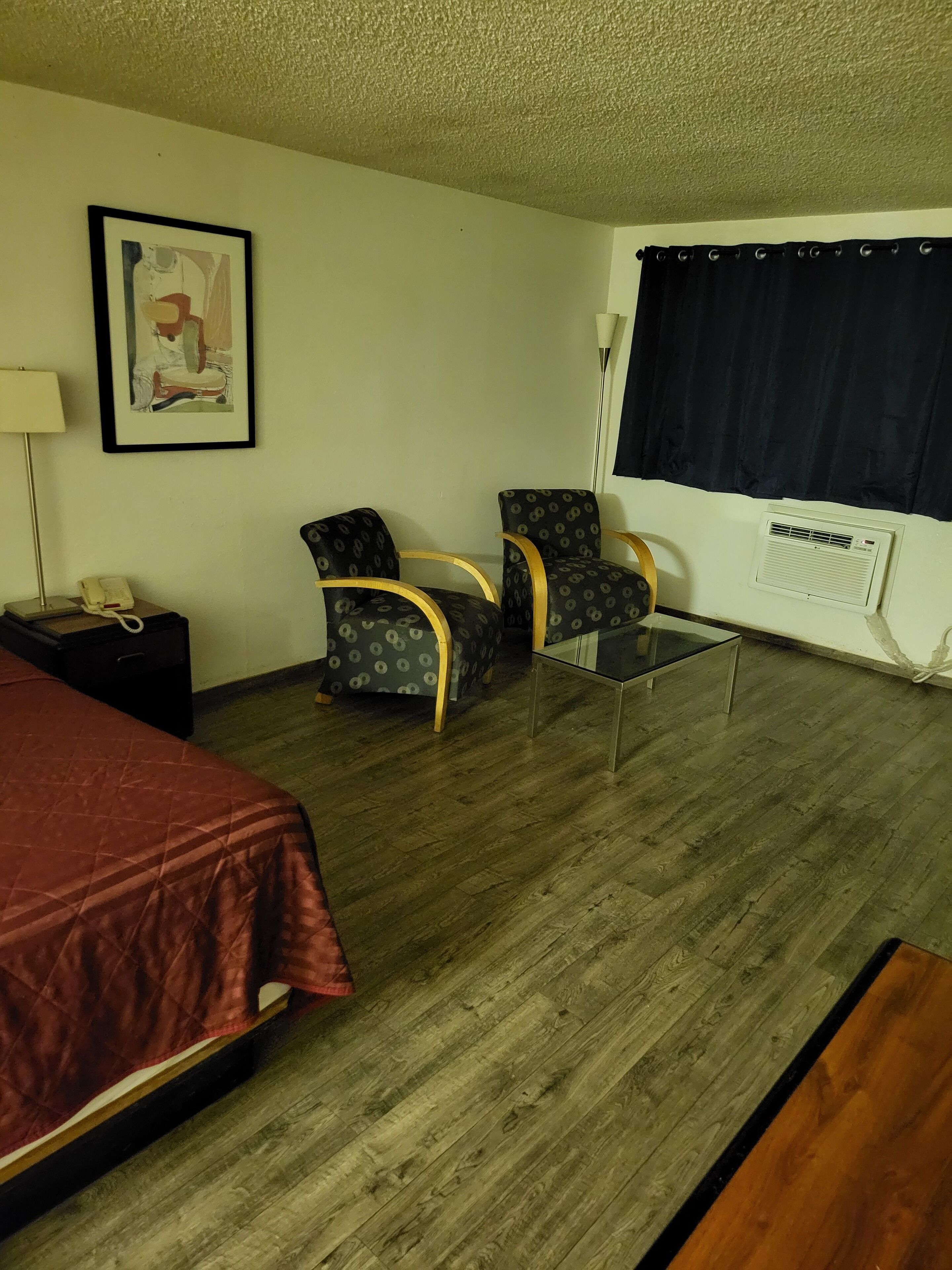 A hotel room with a bed, two armchairs, a small table, a lamp, a phone, a radiator, and a window with curtains.