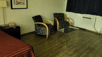 A hotel room with a bed, two armchairs, a small table, a lamp, a phone, a radiator, and a window with curtains.