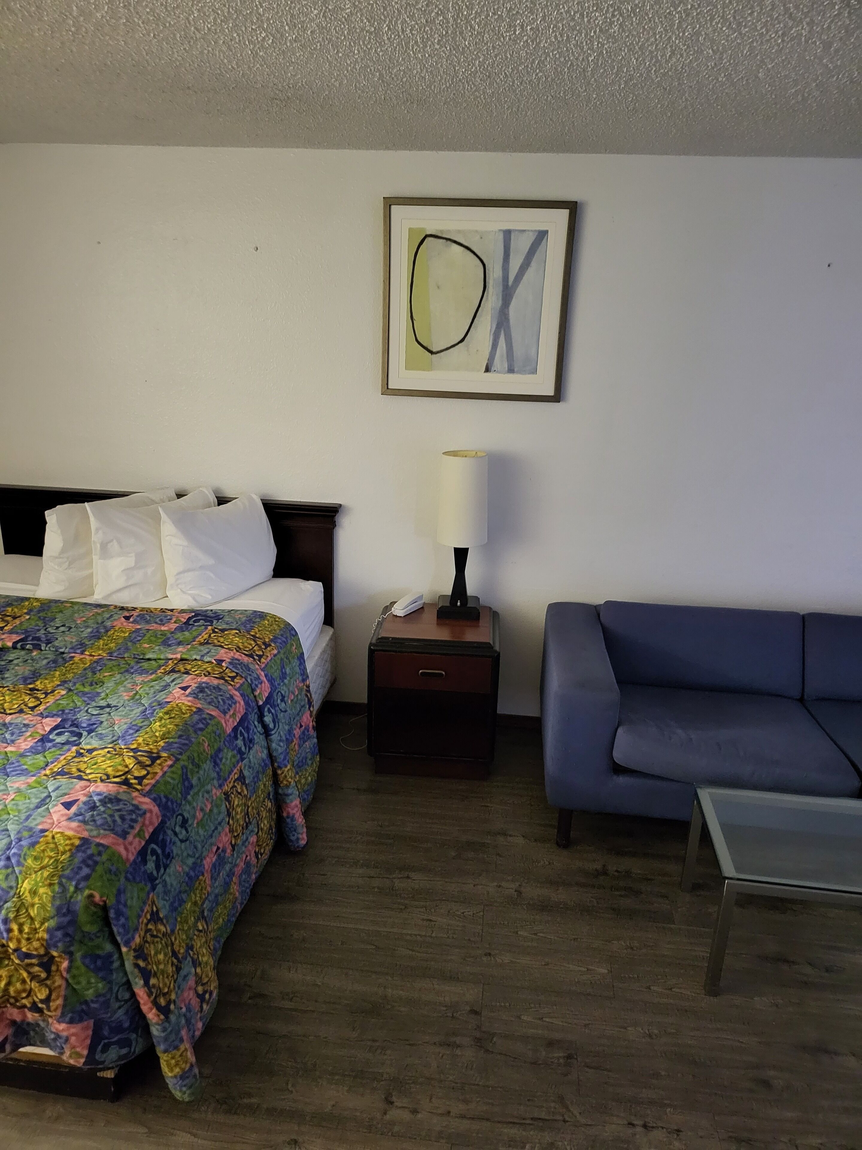 A hotel room with a bed, a bedside table with a lamp, a blue sofa, and a framed abstract painting on the wall.