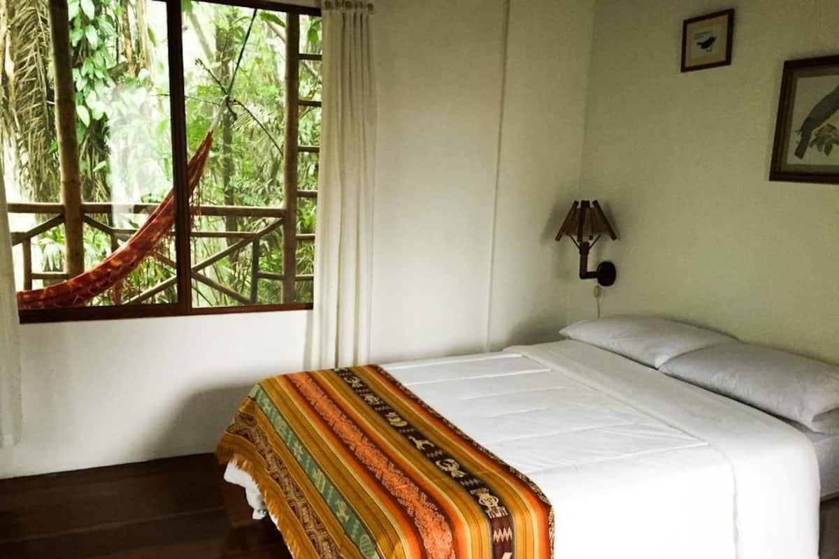 Una habitación con cama, una hamaca, una ventana con cortinas y una lámpara fijada a la pared.