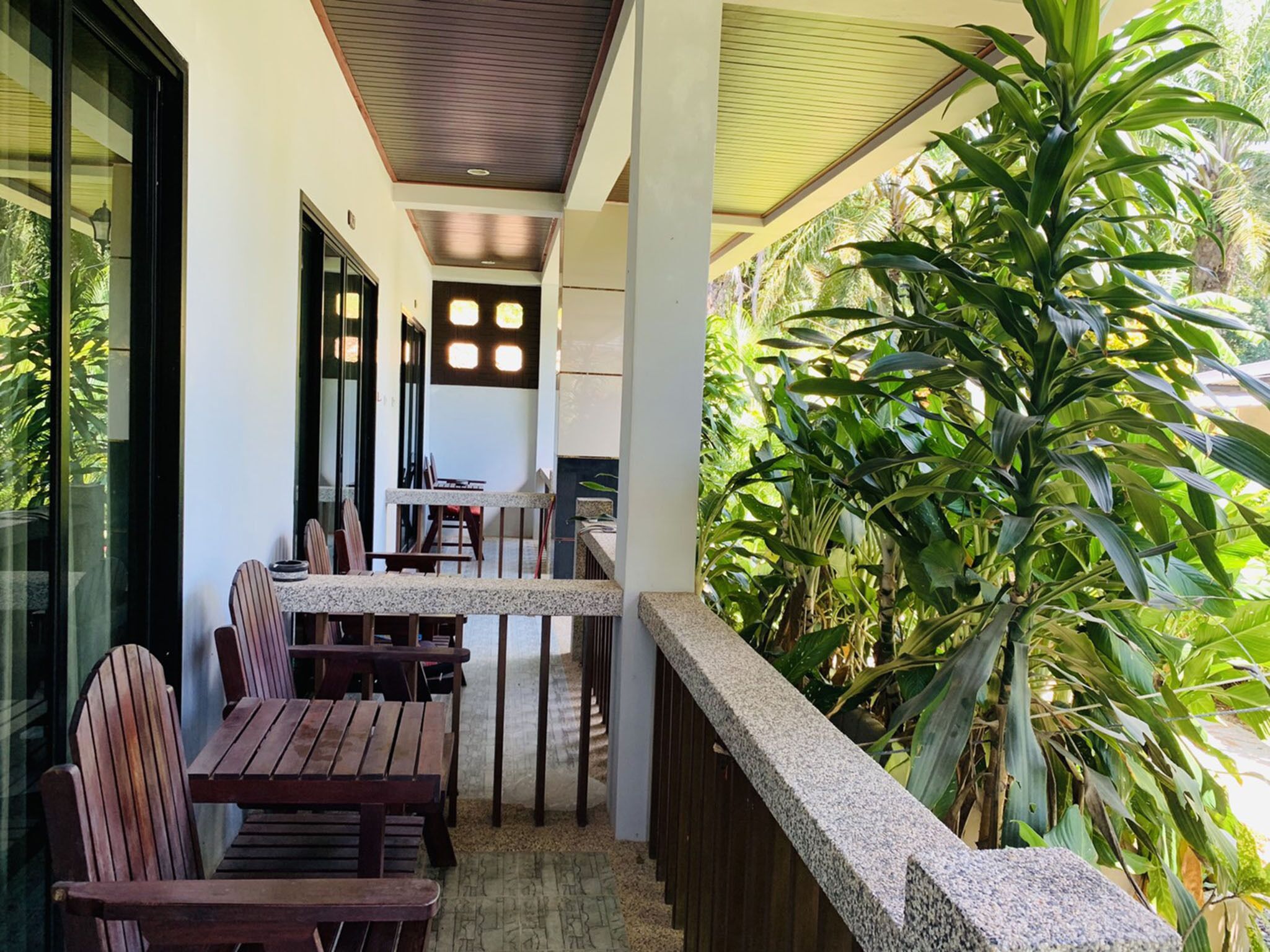 Un espace extérieur aménagé avec une zone de repos couverte, comprenant des tables et des chaises en bois, une balustrade en pierre et une végétation luxuriante.
