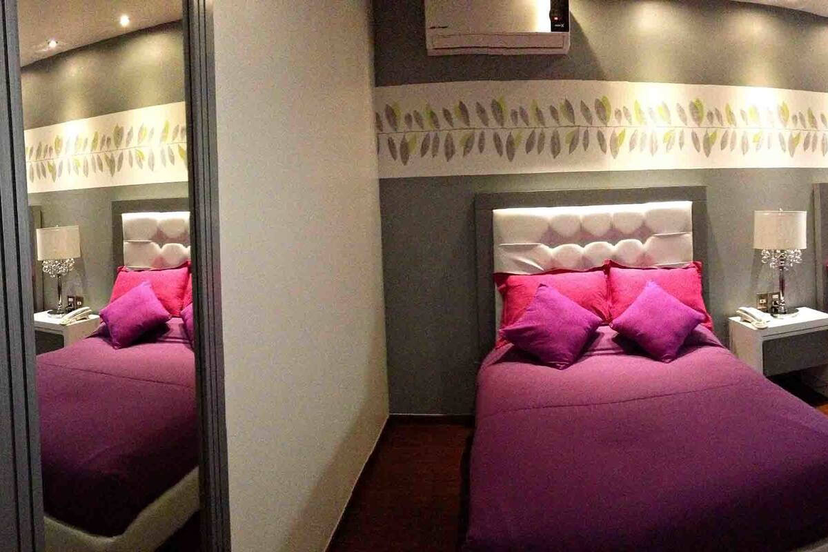 Una habitación de hotel moderna con dos camas, cada una con cabecero capitonado y ropa de cama morada.