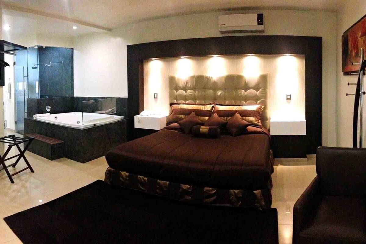 Un dormitorio moderno con una cama grande, una mesita de noche, una silla, un espejo y un baño con lavabo y espejo.