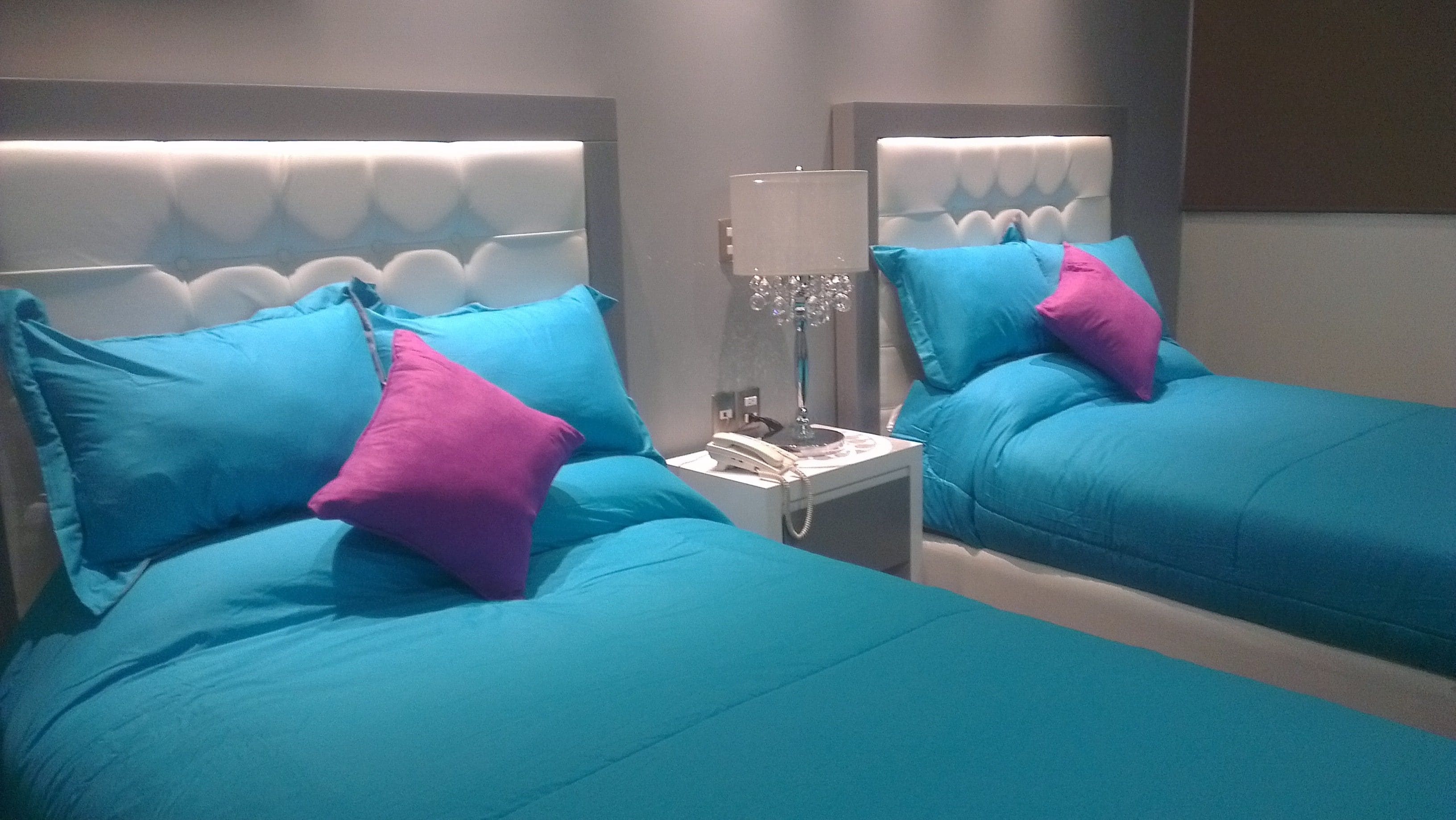 Duas camas com cabeceiras iguais e roupa de cama azul, uma mesinha de cabeceira com abajur e um telefone.