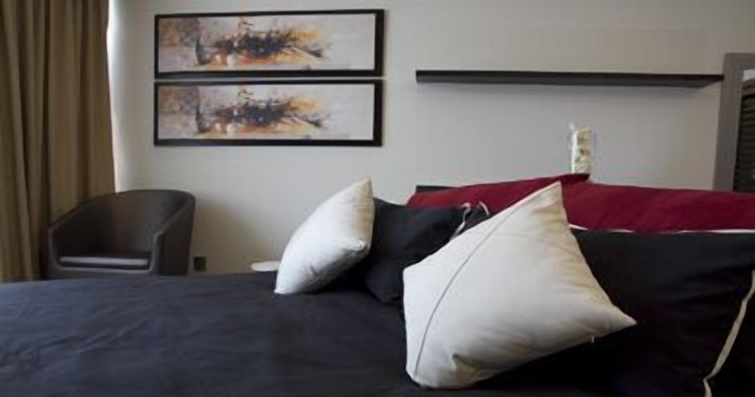 Habitación de hotel con una cama, dos almohadas, una colcha roja y dos cuadros abstractos en la pared.