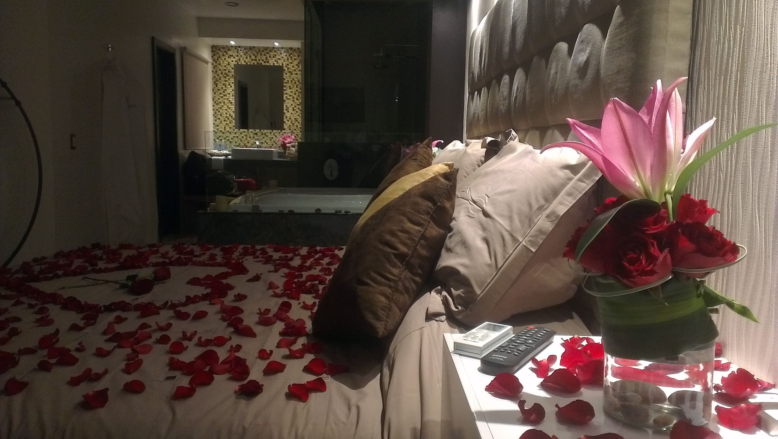 A hotel room with a bed covered in rose petals, a vase with red roses, and a view of a bathroom with a mirror and a sink.
