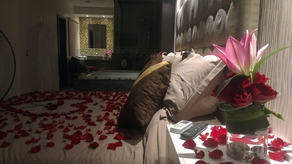 Une chambre d’hôtel avec un lit recouvert de pétales de rose, un vase rempli de roses rouges et une vue sur une salle de bain avec un miroir et un lavabo.