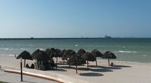 Una playa con sombrillas de paja y un muelle al fondo.