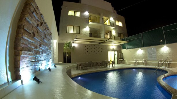 A pool area with a brick wall, a building with multiple balconies, and outdoor seating. - Hotel & Suites Domani (Progreso)