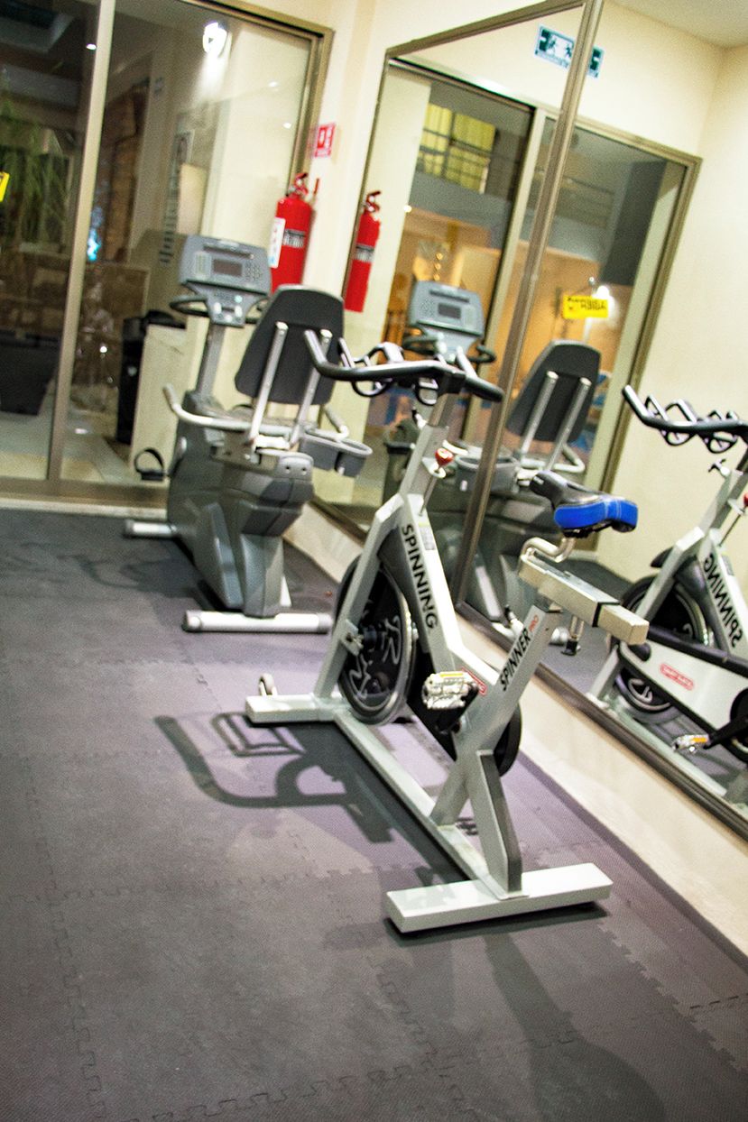 A row of stationary bikes in a gym with a glass wall and a fire extinguisher in the background.