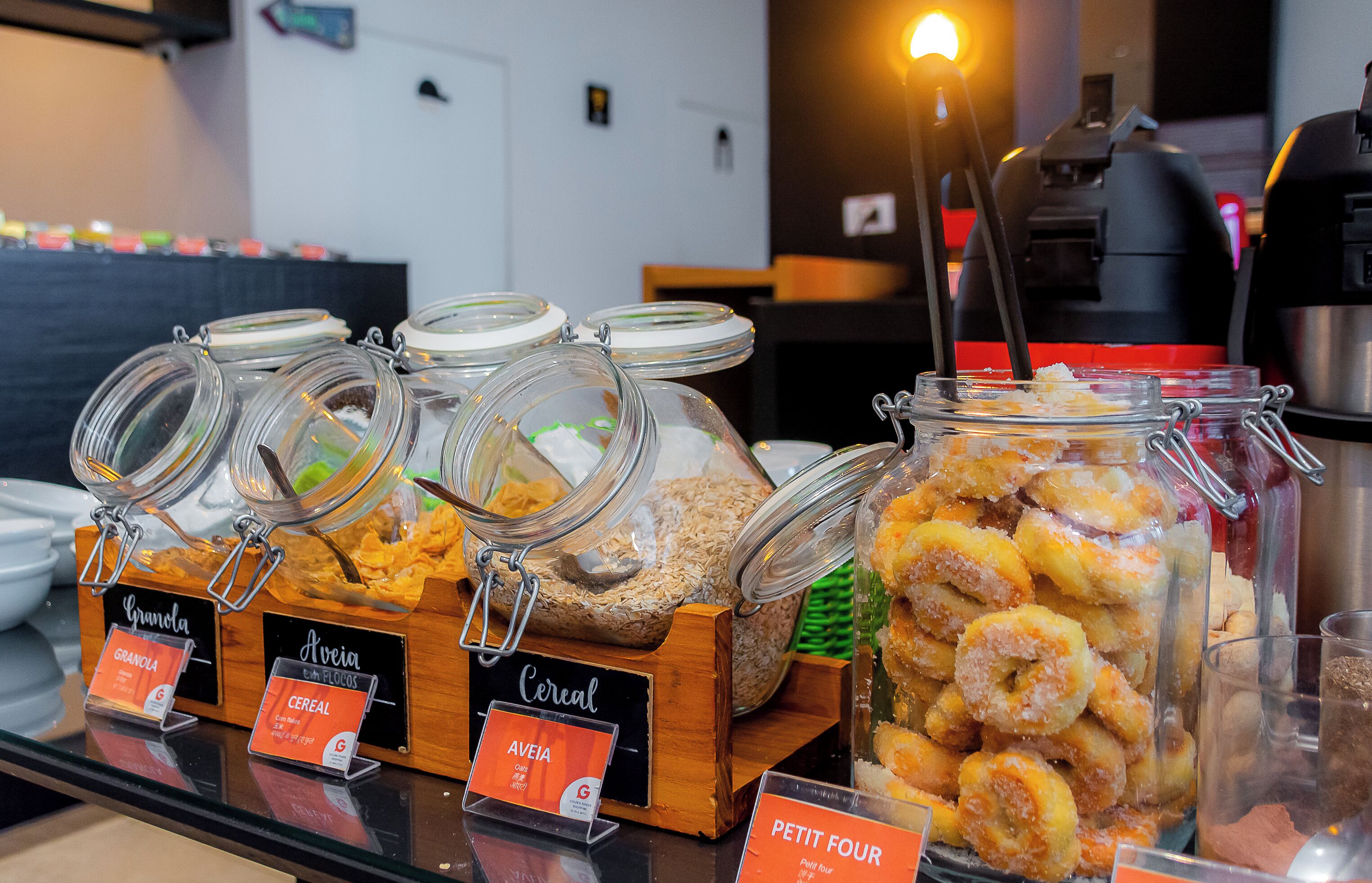 Buffet de café da manhã com itens identificados, incluindo granola, cereais e donuts.
