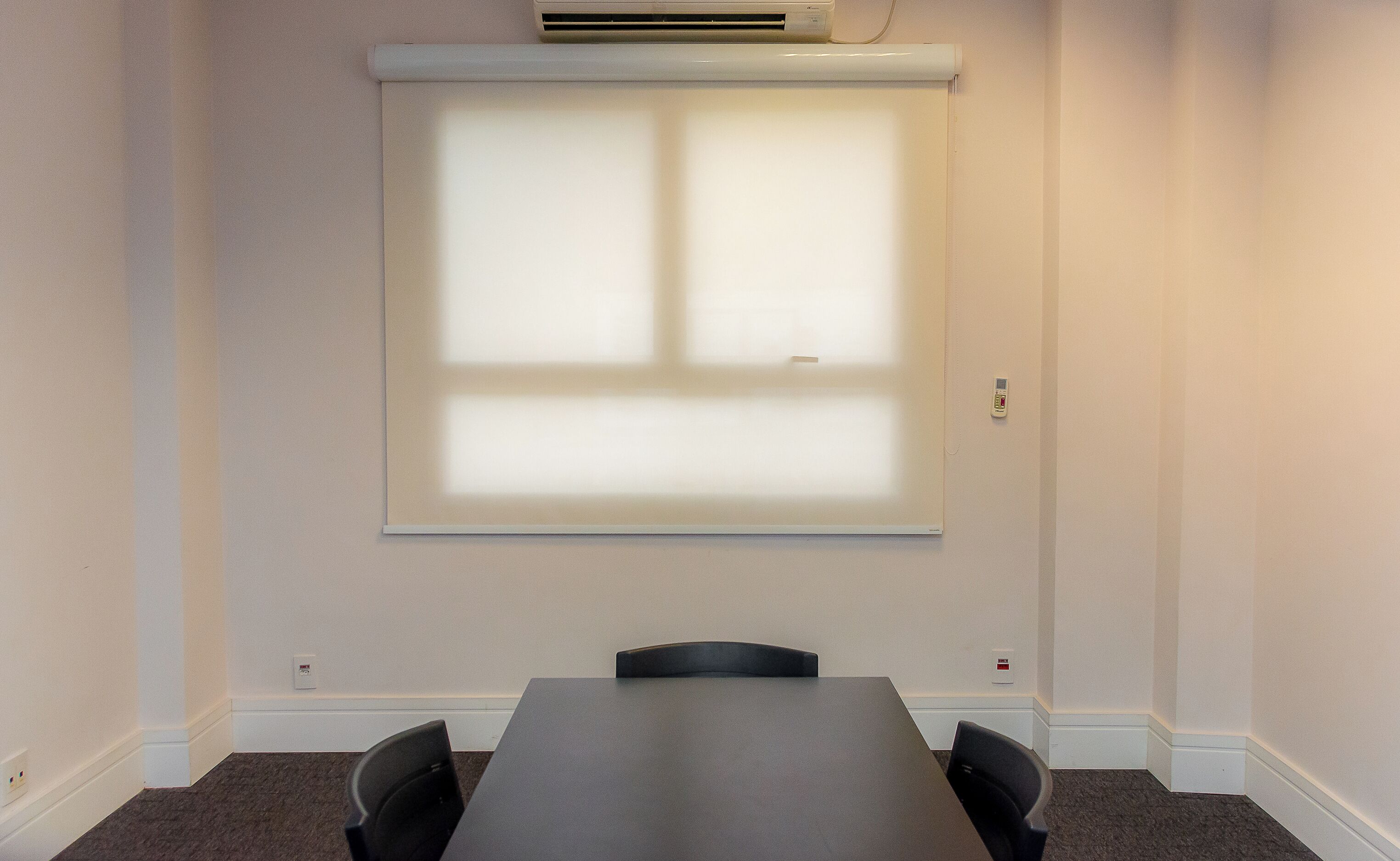 Un salón de conferencias con una gran mesa, cuatro sillas, una pantalla de proyector y un termostato en la pared.