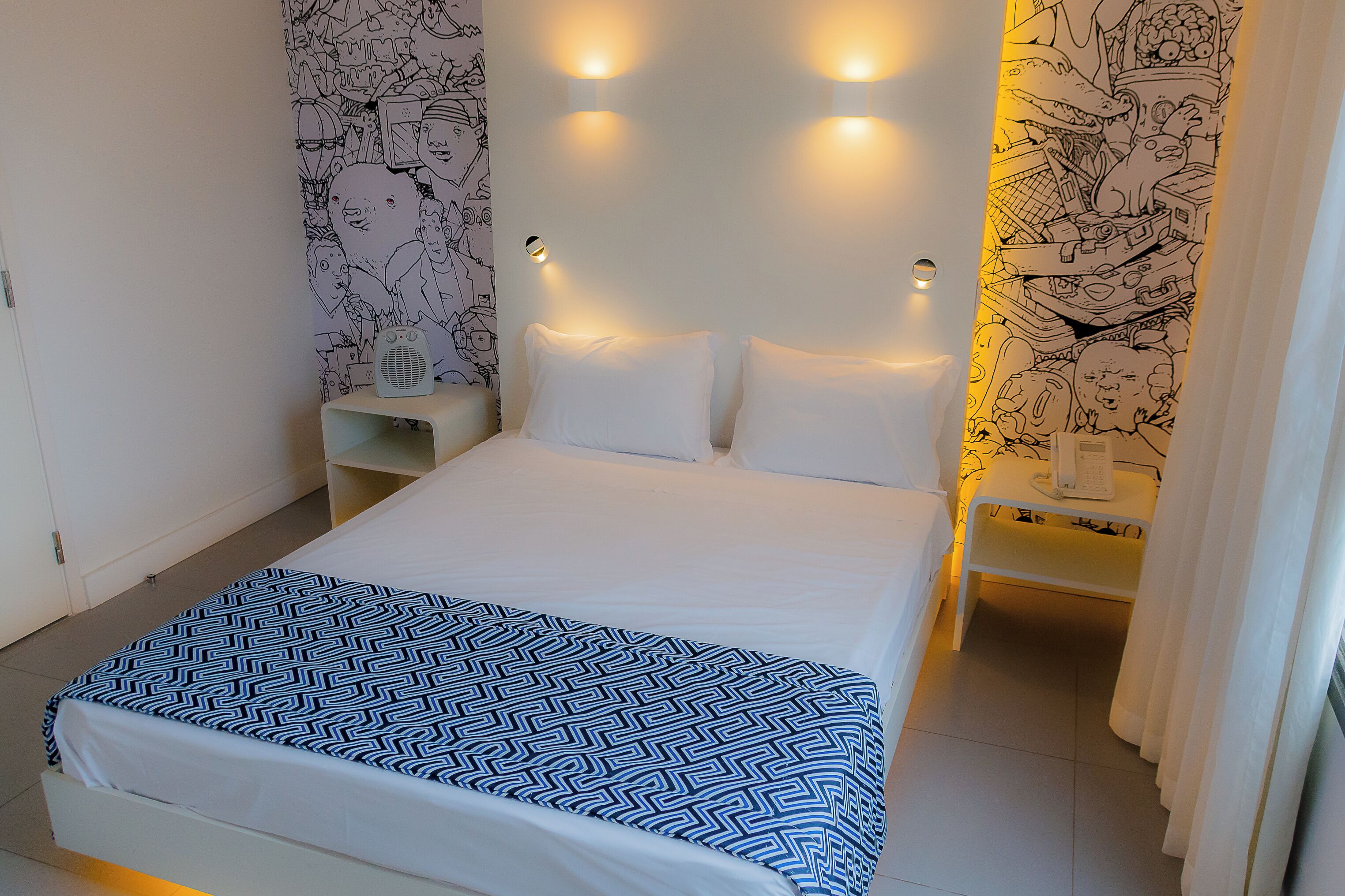 A neatly made bed with a patterned bedspread, a bedside table with a lamp, and a wall with a mural.