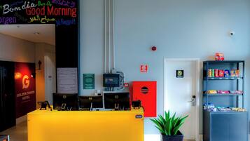 A hotel reception area with a yellow desk, computers, a red safe, and a snack shelf.
