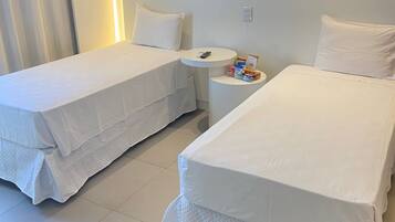 Quarto de hotel com duas camas de solteiro, uma mesa redonda pequena com um controle remoto e uma mesinha de cabeceira com alguns objetos.