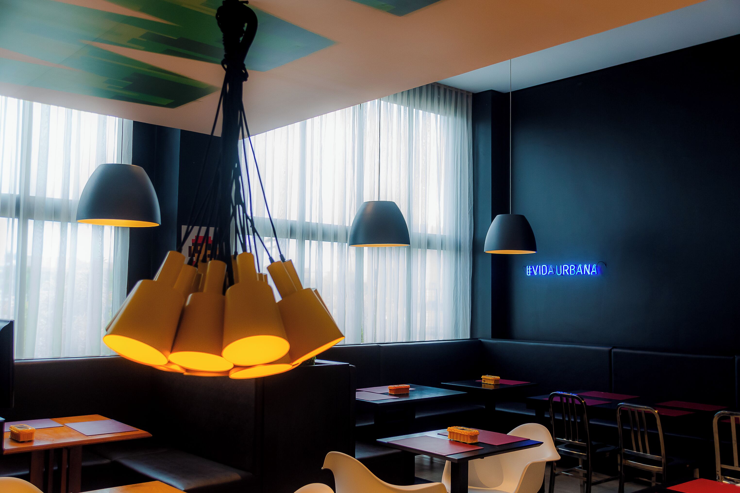 A modern restaurant interior with a dark color scheme, large windows with sheer curtains, and multiple hanging pendant lights.