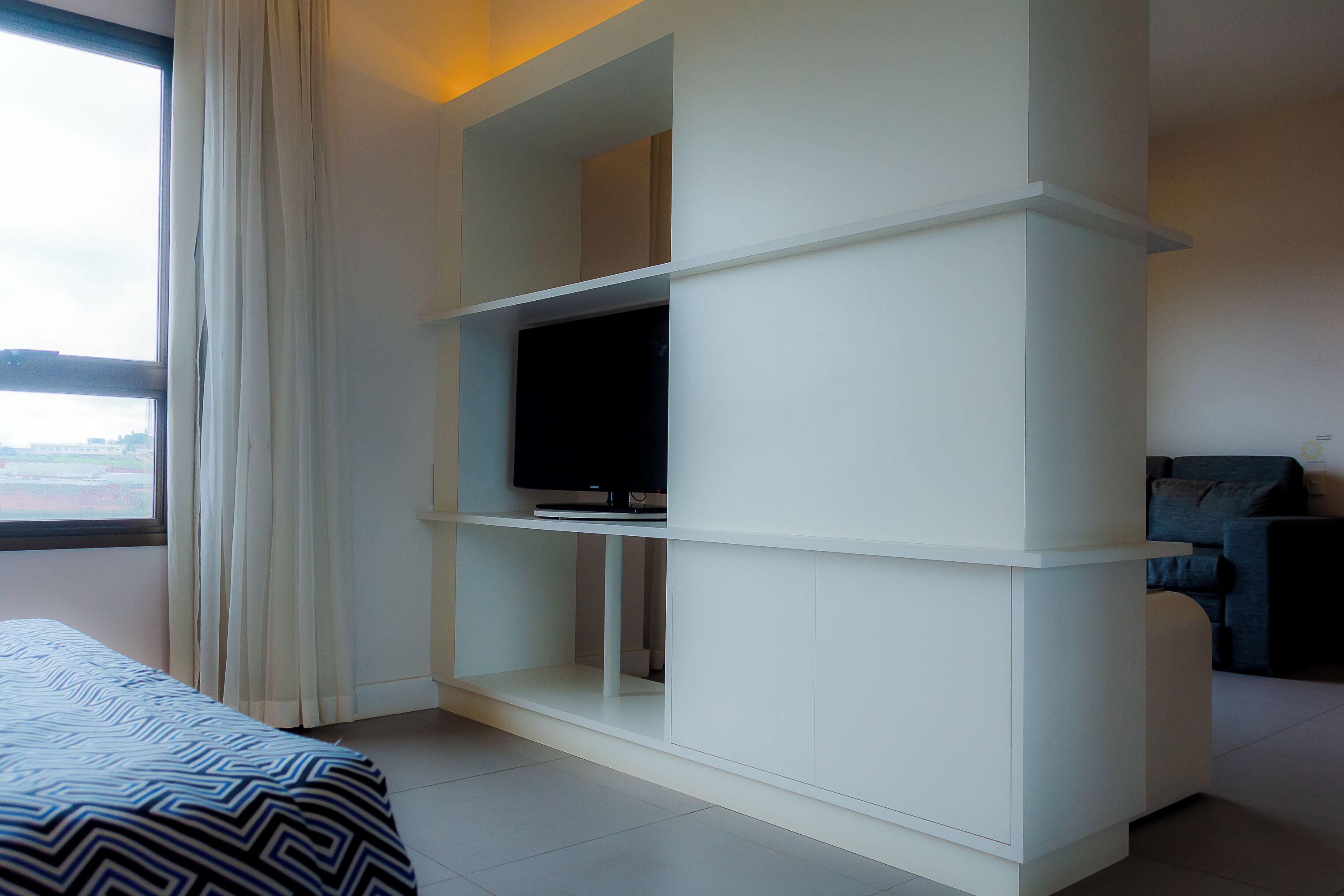 Una habitación de hotel moderna con un ventanal grande, una cama con colcha estampada, un mueble blanco con televisión de pantalla plana y un sofá gris.