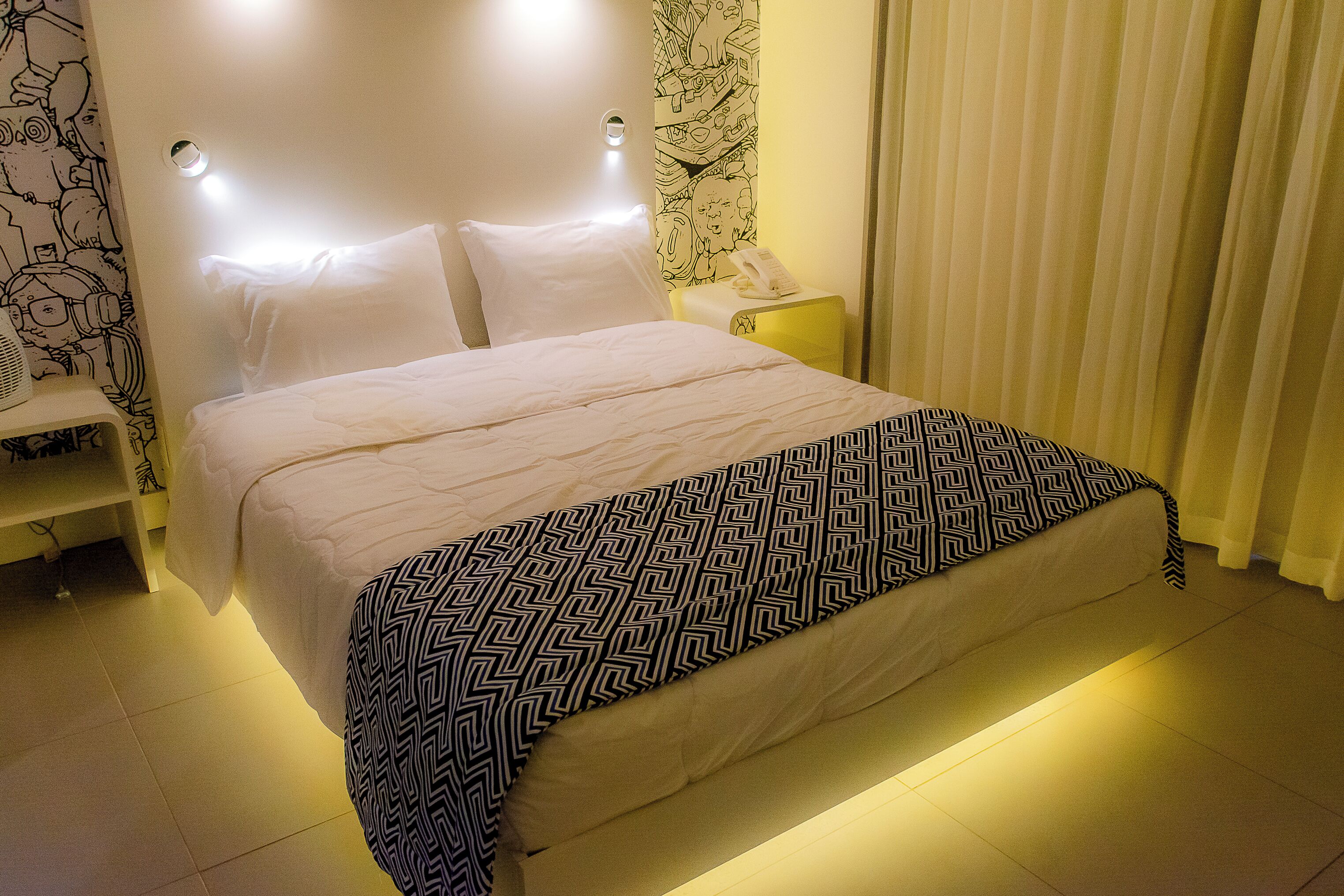 Un lit bien fait, recouvert d’une couvre-lit à motifs, une table de chevet avec une lampe, et un mur tapissé d’un papier peint à motifs.