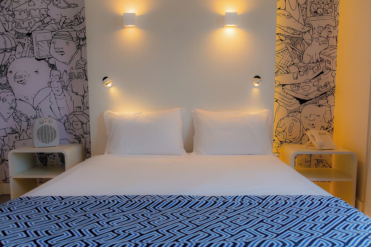 Habitació d'hotel amb un llit, almoinades blanques, un llençol estampat i parets decorades amb il·lustracions en blanc i negre.