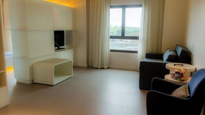 A modern hotel room with a flat-screen TV, a sofa, a chair, a small table, and a window with a view of the city. - Golden Tower Shopping by Fênix Hotéis (Pouso Alegre)