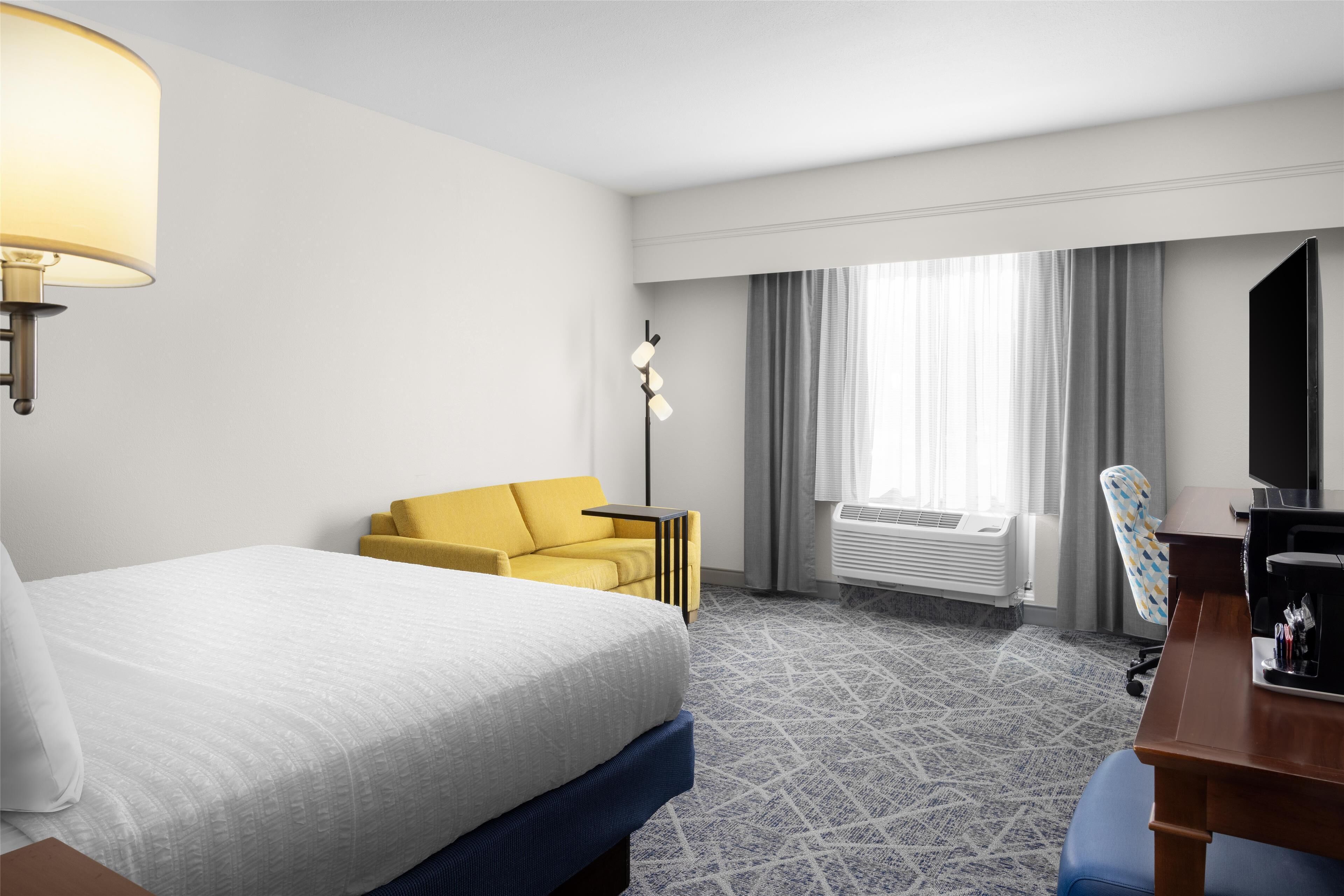 Habitación de hotel con una cama, un sillón amarillo, un escritorio con silla, una lámpara y un ventanal con cortinas.