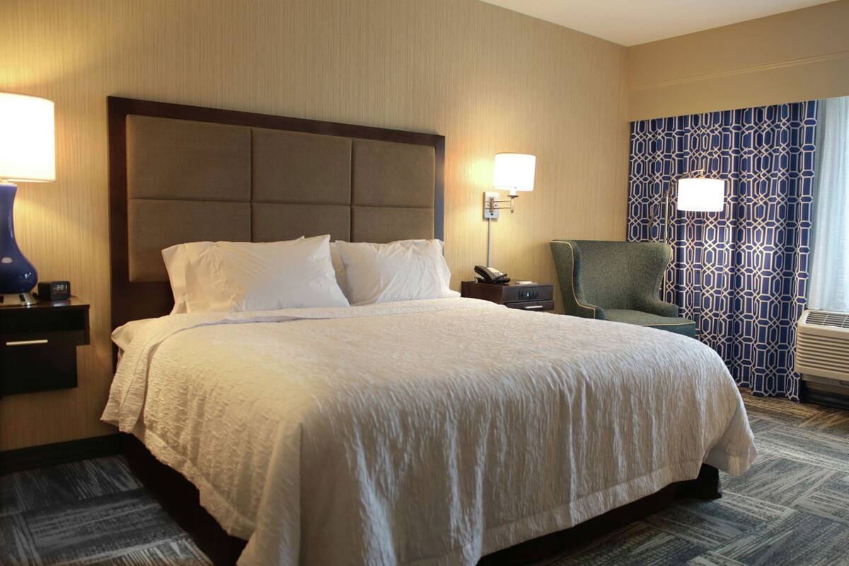 A hotel room with a large bed, a nightstand with a lamp, a chair, and a patterned curtain.