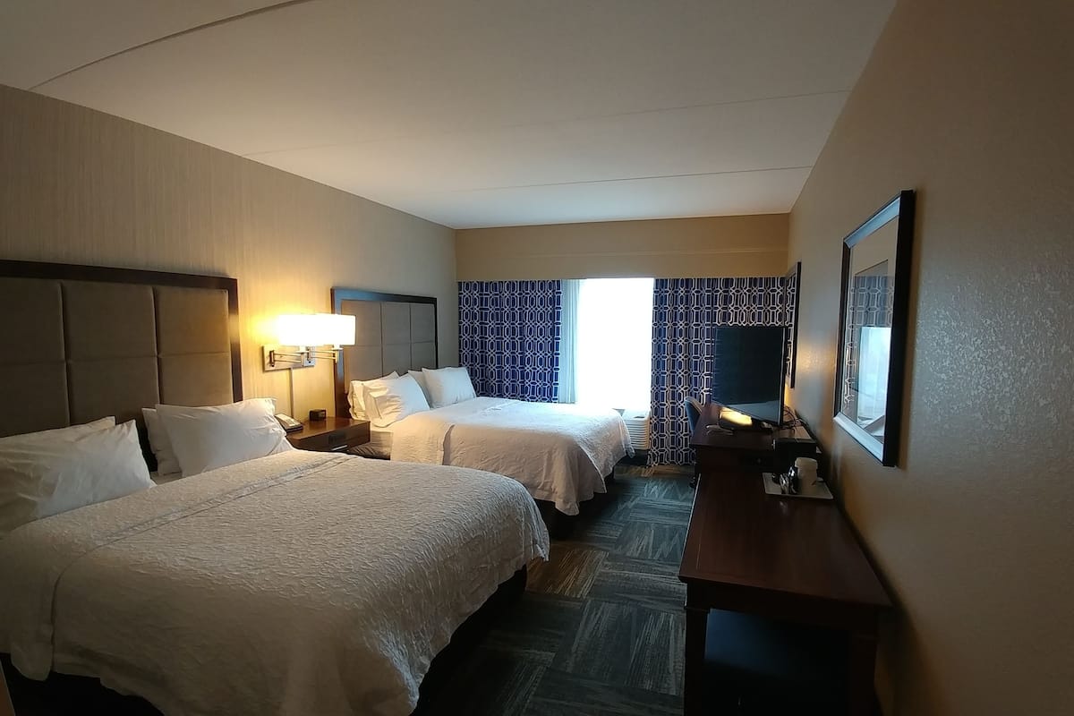 A hotel room with two beds, a desk, a chair, a TV, and a window with patterned curtains.